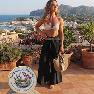 May include: A white bikini top with a center ring detail paired with black high-waisted wrap-style trousers. The outfit is accessorized with a woven straw tote bag featuring a colorful patterned scarf tied to the handle, creating a coordinated look.
