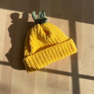 Pode incluir: Um gorro de crochê amarelo com um detalhe de folha verde no topo. O gorro é feito de fio macio e tem um design simples e clássico.