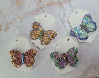 May include: Four white hexagonal gift tags with different colored butterflies. Each butterfly has gold accents and is painted in a realistic style. The tags are tied with white ribbon.