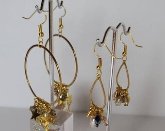May include: Gold-toned hoop and teardrop earrings on display. The hoop earrings feature star charms, while the teardrop earrings have pearl and bead accents. Both sets are displayed on clear acrylic stands.
