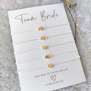 Könnte beinhalten: Fünf weiße Armbänder aus Kordel mit jeweils einem kleinen goldfarbenen Herzanhänger, präsentiert auf einer weißen Karte mit dem Text Team Bride, JGA WIE ER SEIN SOLLTE und EINZIGARTIG, ergänzt durch ein Herzsymbol und einen getrockneten Zweig.