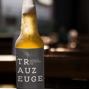 Könnte beinhalten: Eine gekühlte Flasche Bier mit einem schwarzen Etikett, auf dem "TRAUZEUGE" und "MÖCHTEST DU MEIN TRAUZEUGE SEIN?" steht. Die Flasche ist mit Kondensationstropfen bedeckt.