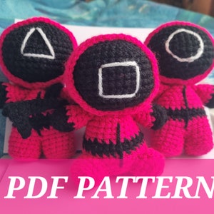 May include: Three pink crocheted plush toys with black details. Each toy has a different geometric shape on its head: a triangle, a square, and a circle. The toys are sitting on a white surface with the text "PDF PATTERN" in pink letters.