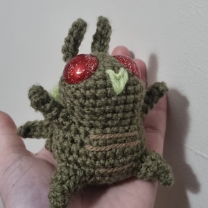 May include: A green crocheted plush toy with red glitter eyes and a green heart on its chest. The toy is sitting on a hand.