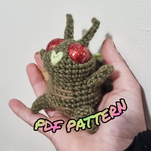 May include: A crocheted green and brown plush toy with red glitter eyes. The toy is being held in a person's hand. The text "PDF PATTERN" is written in rainbow colors on the hand.