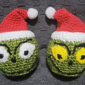 May include: Two crocheted Grinch ornaments wearing red and white Santa hats. The Grinch's face is green with yellow eyes and a black frown.