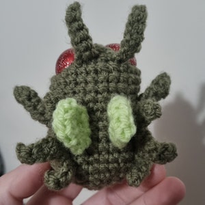 May include: A crocheted green bug with red eyes and light green wings. The bug is sitting in a hand.