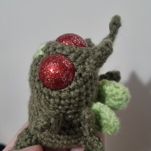 May include: A green crocheted plush toy with red glitter eyes and green crocheted arms and legs.