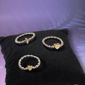May include: Three stretch rings made of alternating silver and gold-toned beads. One ring features a gold-toned cross, one features a plain gold-toned heart, and one features a gold-toned heart charm engraved with the word mama.