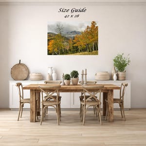 Mockup of a 42 by 28 inch autumn mountain landscape print displayed on a dining room wall above a wooden table.