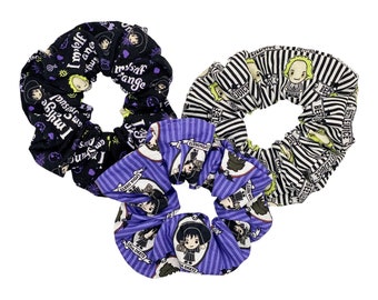Puede incluir: Conjunto de tres scrunchies negros, blancos y morados con un patrón repetido de un personaje de dibujos animados con el texto "I myself am strange" y "Beetlejuice" en los scrunchies.