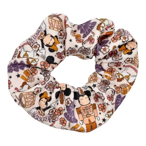 May include: A white scrunchie with a whimsical print featuring Mickey Mouse, Minnie Mouse, and other characters. The print includes a carousel, stars, and flowers in shades of pink, purple, and brown.