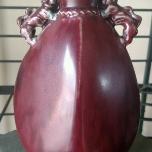 May include: A dark red ceramic vase with two lion-shaped handles. The vase has a textured surface and a rounded body.