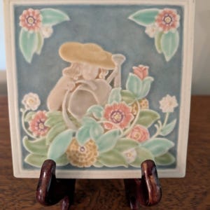May include: Square ceramic tile with a relief design of a figure in a hat, surrounded by flowers and foliage in pastel colors. The tile is mounted on a dark wooden stand, showcasing the artwork.