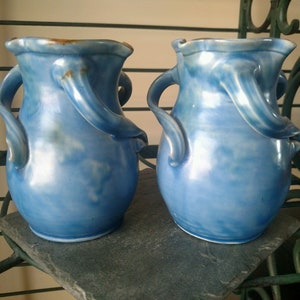 May include: Two blue ceramic vases with unique, sculpted handles. The vases have a light blue glaze with a subtle iridescent sheen.