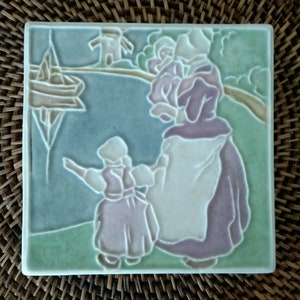 May include: Square ceramic tile with a bas-relief scene of a mother and child near a body of water. The tile features a windmill, boat, and figures in pastel colors of green, blue, and lavender. The tile is likely a decorative piece.