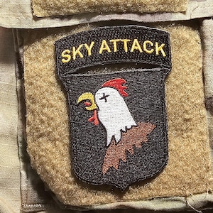 May include: A black patch with the words "SKY ATTACK" in yellow. The patch features a white chicken head with a red comb and a yellow beak. The patch is on a tan background.
