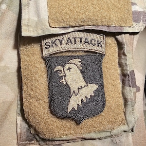 May include: A military patch with the text "SKY ATTACK" above a black and gold emblem featuring a bird of prey. The patch is attached to a tan fabric with a camouflage pattern.