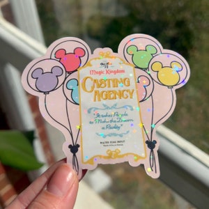 May include: A pink and white sticker with a glittery finish featuring a sign that reads "Magic Kingdom Casting Agency" with a quote from Walter Elias Disney. The sign is surrounded by five colorful balloons with Mickey Mouse ears.