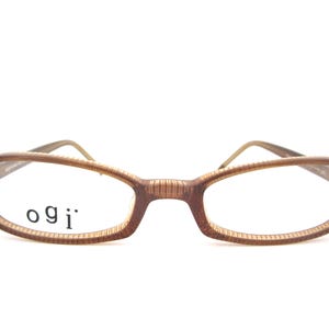 Pode incluir: Óculos retangulares castanhos com o nome da marca "ogi" impresso na lente. As armações têm um aspeto de grão de madeira. As hastes são de cor castanho claro.