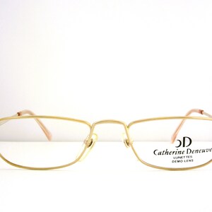 Puede incluir: Gafas de oro con montura rectangular y detalles azules. Las gafas tienen una etiqueta "Catherine Deneuve Lunettes Demo Lens" en la patilla derecha.