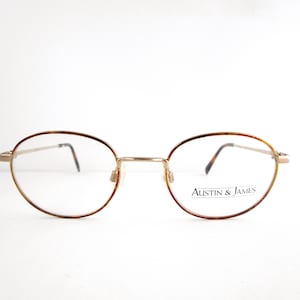 May include: Oval-shaped eyeglasses with a gold-colored frame and tortoise shell accents on the upper rims. The lenses are clear, and the brand name "Austin & James" is printed on the right lens.