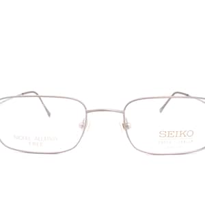 Op de afbeelding: Zilverkleurige rechthoekige bril met dunne metalen monturen. Het linkerglas bevat de tekst SEIKO SUPER TITANIUM en het rechterglas bevat de tekst NICKEL ALLERGY FREE. De bril heeft een neusbrug, dunne pootjes en neuspads.