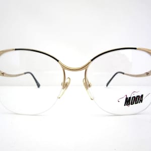 May include: Gold-coloured eyeglasses with a black accent along the top rim and temples. The lenses are clear, and the frame features a delicate design. The word "MODA" is printed on the right lens.