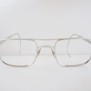 May include: Silver-toned eyeglasses with a double bridge and rectangular lenses. The frames have a classic aviator style, with a thin metal construction. The glasses are set against a plain white background, highlighting their minimalist design.