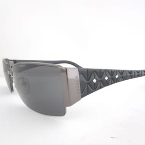 May include: Dark grey sunglasses with a geometric pattern on the temples. The sunglasses have a rectangular shape with dark grey lenses and a light grey frame. The brand name "ESCADA" is visible on the temple.