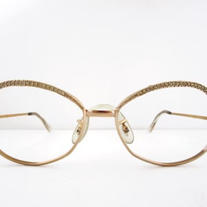 Könnte beinhalten: Goldfarbene Brille mit ovalem Design. Die oberen Ränder sind mit kleinen, funkelnden Details verziert. Die Rahmen haben ein zartes, drahtartiges Aussehen, und die Gläser sind klar. Der Nasensteg und die Bügel sind ebenfalls goldfarben.