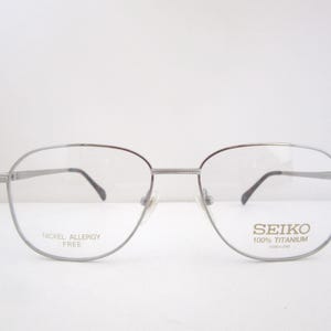 May include: Silver-toned Seiko eyeglasses with a rounded rectangular frame. The lenses are clear, and the temples are thin. The text "SEIKO 100% TITANIUM" and "NICKEL ALLERGY FREE" are visible on the lenses.