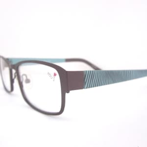 May include: Rectangular eyeglasses with a matte dark brown frame and clear lenses. The temples feature a teal and dark brown striped pattern. The right lens has the text "YOU'S" printed in black with a small red tulip icon above the letter S.