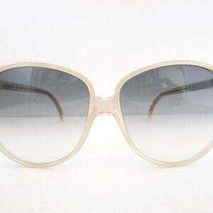 May include: Large, pale beige sunglasses with gradient lenses. The lenses transition from dark grey to a lighter shade. The frames are a light colour, and the temples are thin and brown. A classic accessory, suitable for sunny days.