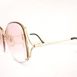 May include: Vintage oversized sunglasses with a gold metal frame and pink gradient lenses. The glasses have a unique shape with a thick frame around the lenses and black temple tips. The design is reminiscent of the 1970s.