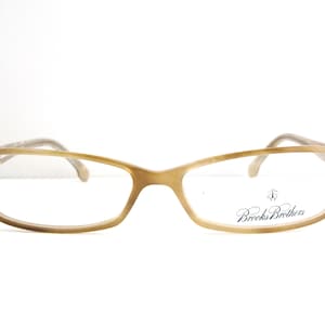 May include: Tan rectangular eyeglasses with a subtle cat-eye shape. The frames are a light brown color, with the brand name "Brooks Brothers" printed on the right lens. The temples are a lighter shade of brown.