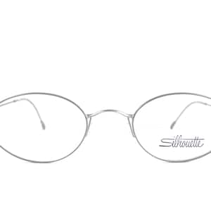 May include: Silver-toned Silhouette eyeglasses with oval lenses. The glasses have a thin, minimalist frame and the brand name "Silhouette" is printed on the right lens. The design is sleek and modern.