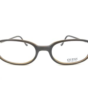 Puede incluir: Gafas de forma ovalada con montura marrón oscuro y borde interior marrón claro. La marca "GUESS" está impresa en la lente inferior derecha. Las gafas tienen un diseño clásico y discreto.