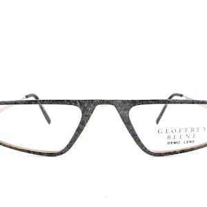 Może przedstawiać: Okulary Geoffrey Beene o unikalnym, kanciastym designie. Oprawki są szare w cętki ze złotą wewnętrzną obwódką. Tekst "GEOFFREY BEENE DEMO LENS" jest wydrukowany na prawej soczewce.