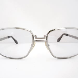 May include: Silver-framed eyeglasses with a square shape. The glasses have clear lenses and dark temples. The bridge of the glasses is a straight bar. The glasses are set against a white background.
