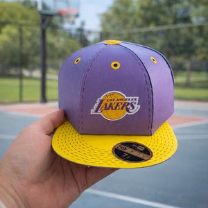 May include: A purple and yellow Los Angeles Lakers baseball cap. The cap has the team's logo on the front and a yellow peak. The cap is a 59FIFTY style.