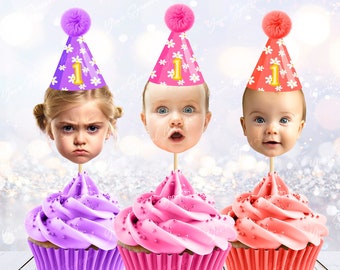 May include: Three cupcakes with pink and purple frosting, topped with a baby's face wearing a purple, pink, or red party hat with a white flower pattern and the number "1" on it.