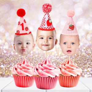 May include: Three pink cupcakes with pink frosting and red cupcake liners. Each cupcake has a red, white, and pink birthday hat with a baby's face on top. The hats have strawberries and the number "1" on them.