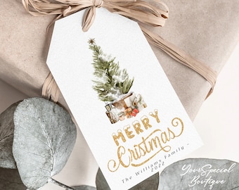 May include: A white gift tag with a watercolor illustration of a Christmas tree and presents. The tag reads "MERRY Christmas" in gold glitter lettering, with the family name and year below. The tag is tied to a wrapped gift with a raffia bow.