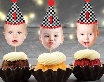 May include: Three cupcake toppers featuring a baby's face with a red and black checkered racing hat. The hats have a red pom-pom and the number "1" on the front.