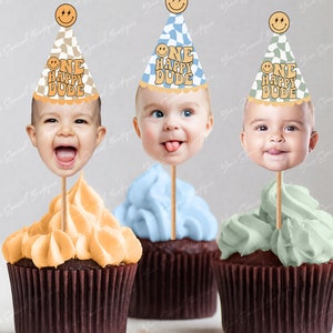 May include: Three cupcakes with different coloured frosting, each topped with a cake topper featuring a baby's face wearing a party hat with the text "One Happy Dude".