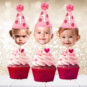 Puede incluir: Tres toppers de cupcake rosa y blanco con un sombrero de fresa rosa y blanco con el número 1. Cada topper tiene la cara de un niño diferente. Los cupcakes son rojos y rosas.