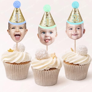 Könnte beinhalten: Drei Cupcakes mit weißem Zuckerguss, jeweils gekrönt mit einem Babygesicht, das einen goldenen Glitzer-Partyhut trägt. Die Hüte haben die Zahlen 1 und 2. Jeder Cupcake hat ein dekoratives Element, darunter ein Stern und eine Kokosnusskugel.