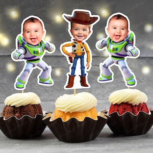 May include: Cupcakes with toppers featuring a baby's face as Buzz Lightyear and Woody from Toy Story. The cupcakes have white frosting and are in black paper cups. The background is a blurred gray.
