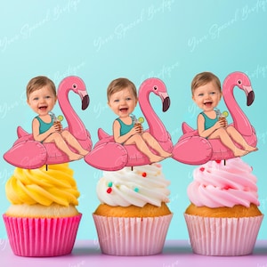Op de afbeelding: Drie cupcakes met roze, witte en gele glazuur, elk bekroond met een flamingo-vormige decoratie en een cartoon baby-afbeelding. De cupcakes staan in roze papieren cups tegen een lichtblauwe achtergrond.